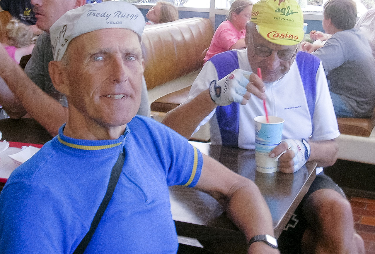 Jobst Brandt and a friend sit at a table in an ice cream parlor. Jobst wears a blue polypropylene bike jersey with yellow trim and a Fredy Ruegg Velos cycling cap, while his friend, who is looking into a paper cup, wears a white and purple bicycle jersey and a yellow ag2 cycling cap.