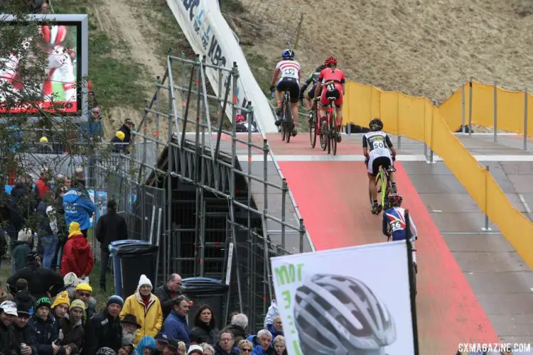 Riders in the field head over the flyover while the big screen shows the front of the race. 2017 World Cup Koksijde. © B. Hazen / Cyclocross Magazine